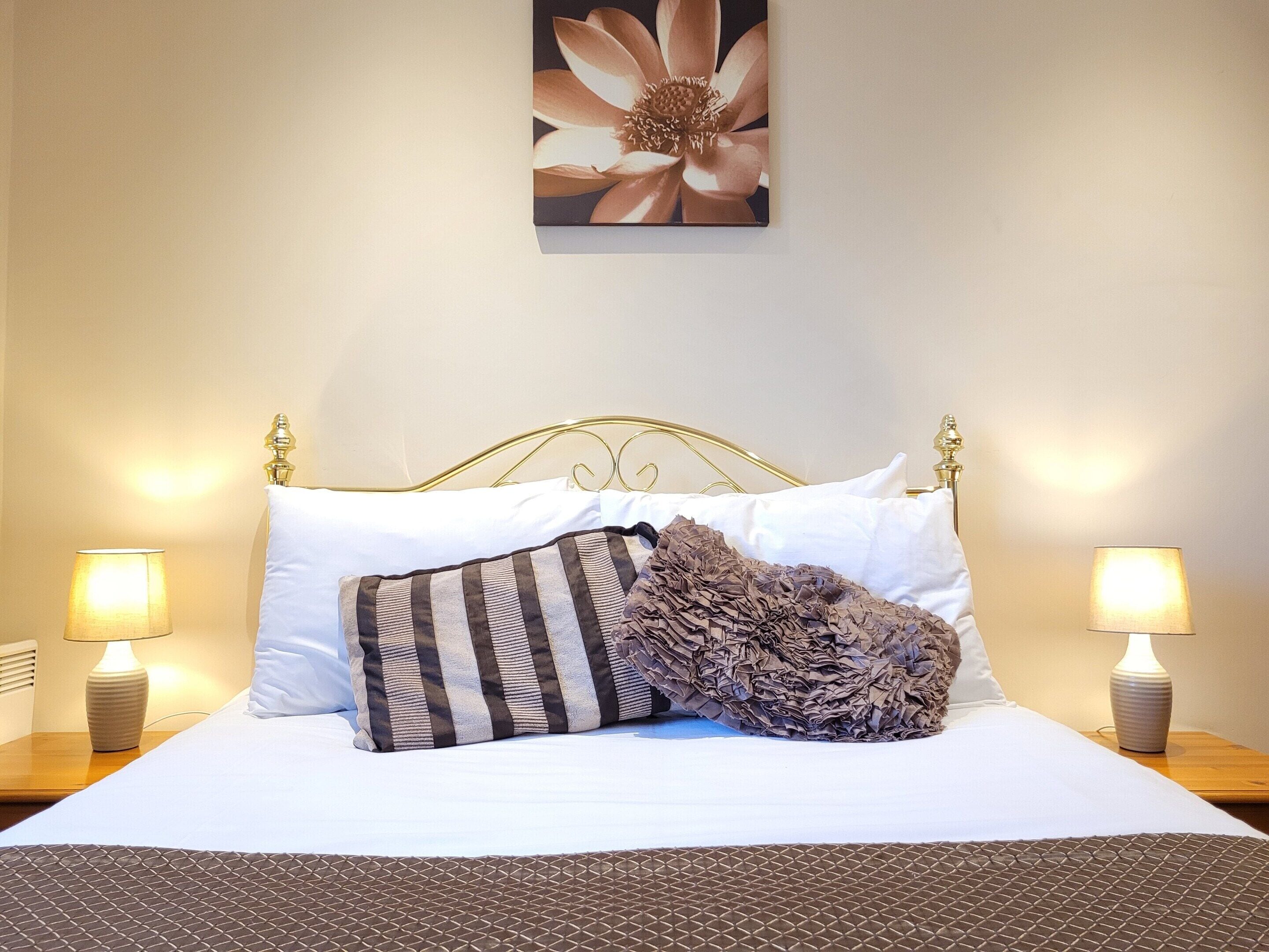 Una cama bien hecha con sábanas blancas, un almohadón a rayas y una colcha texturizada. Hay dos lámparas de noche sobre mesitas de madera, y una obra de arte floral cuelga sobre la cama.
