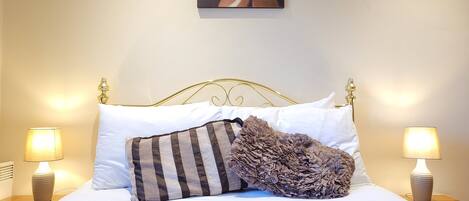 Una cama bien hecha con sábanas blancas, un almohadón a rayas y una colcha texturizada. Hay dos lámparas de noche sobre mesitas de madera, y una obra de arte floral cuelga sobre la cama.