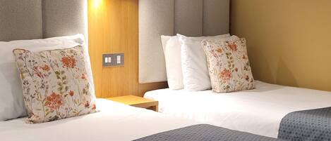 Habitación de hotel con dos camas, cada una con ropa blanca y almohadas de estampado floral. Tiene cabecera de madera con iluminación integrada y una mesita de noche pequeña.