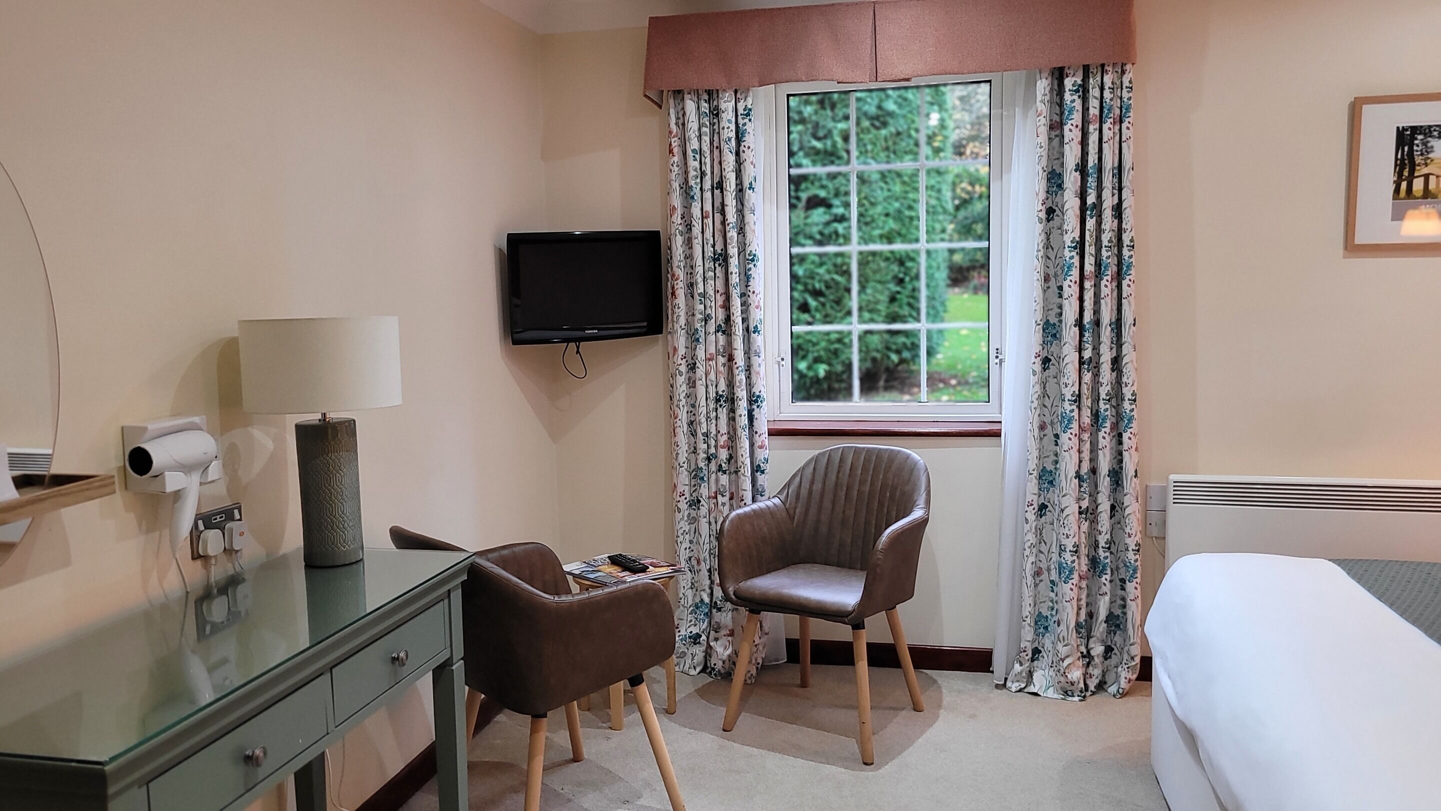 A hotel room with a bed, a desk with a lamp, a chair, a TV, and a window with curtains.