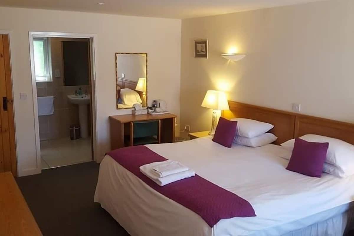 A hotel room with a bed, a bedside table, a lamp, a mirror, and a bathroom visible through an open door.