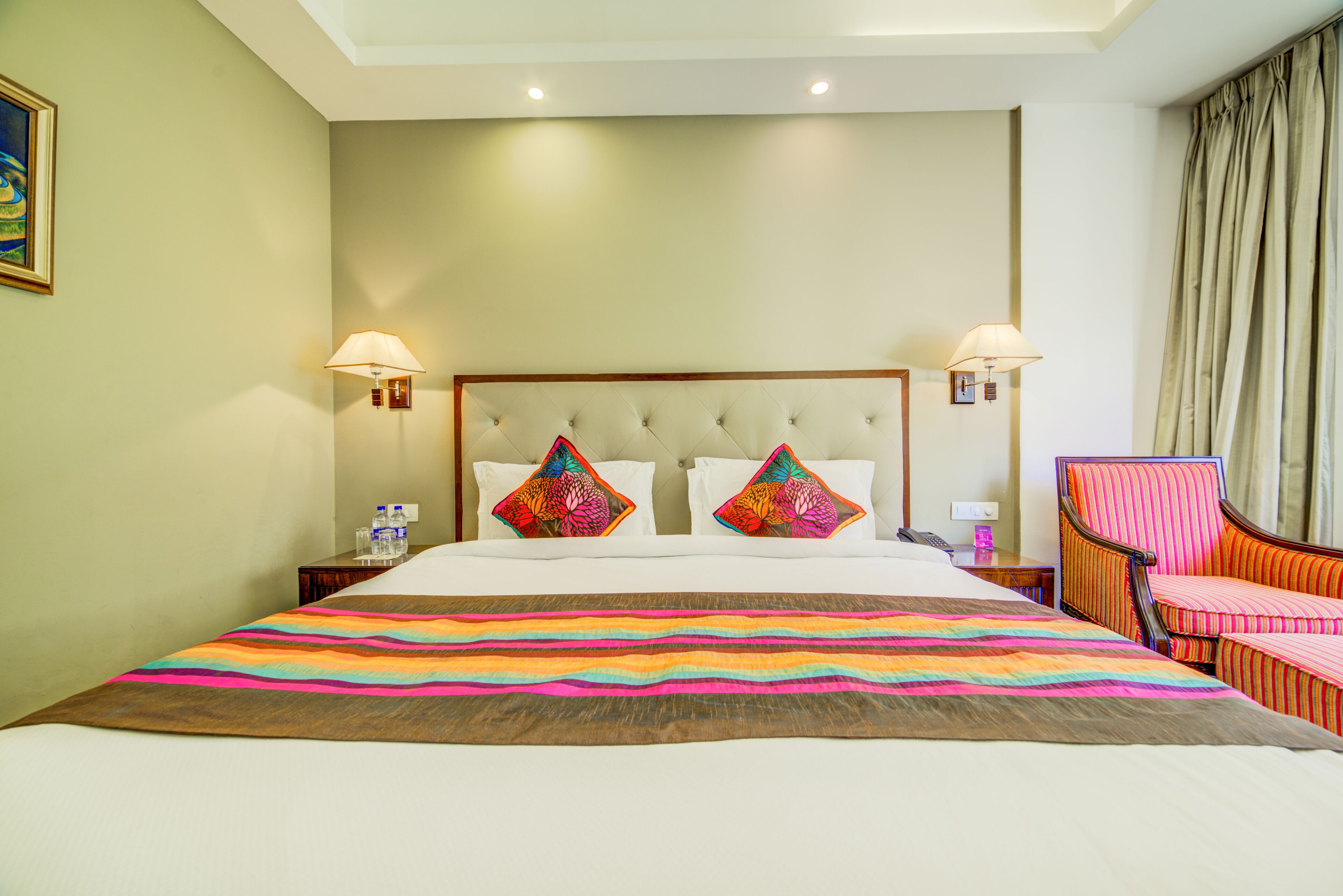 A hotel room with a large bed, two decorative pillows, a striped chair, and a nightstand with a lamp.