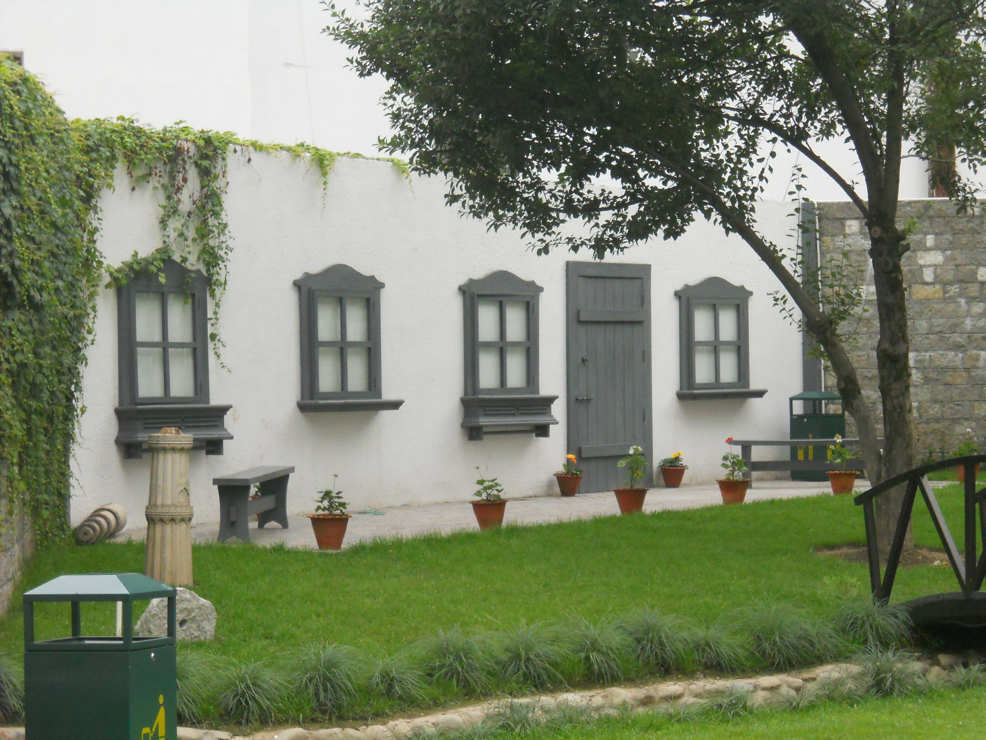 A building with three windows and a door, surrounded by a grassy area with potted plants.