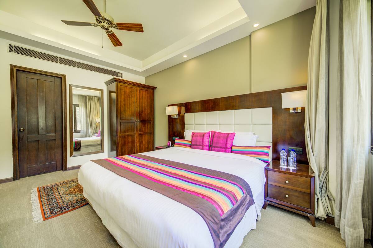 Une chambre d’hôtel avec un grand lit, une tête de lit en bois, une table de chevet avec deux bouteilles et un ventilateur de plafond.