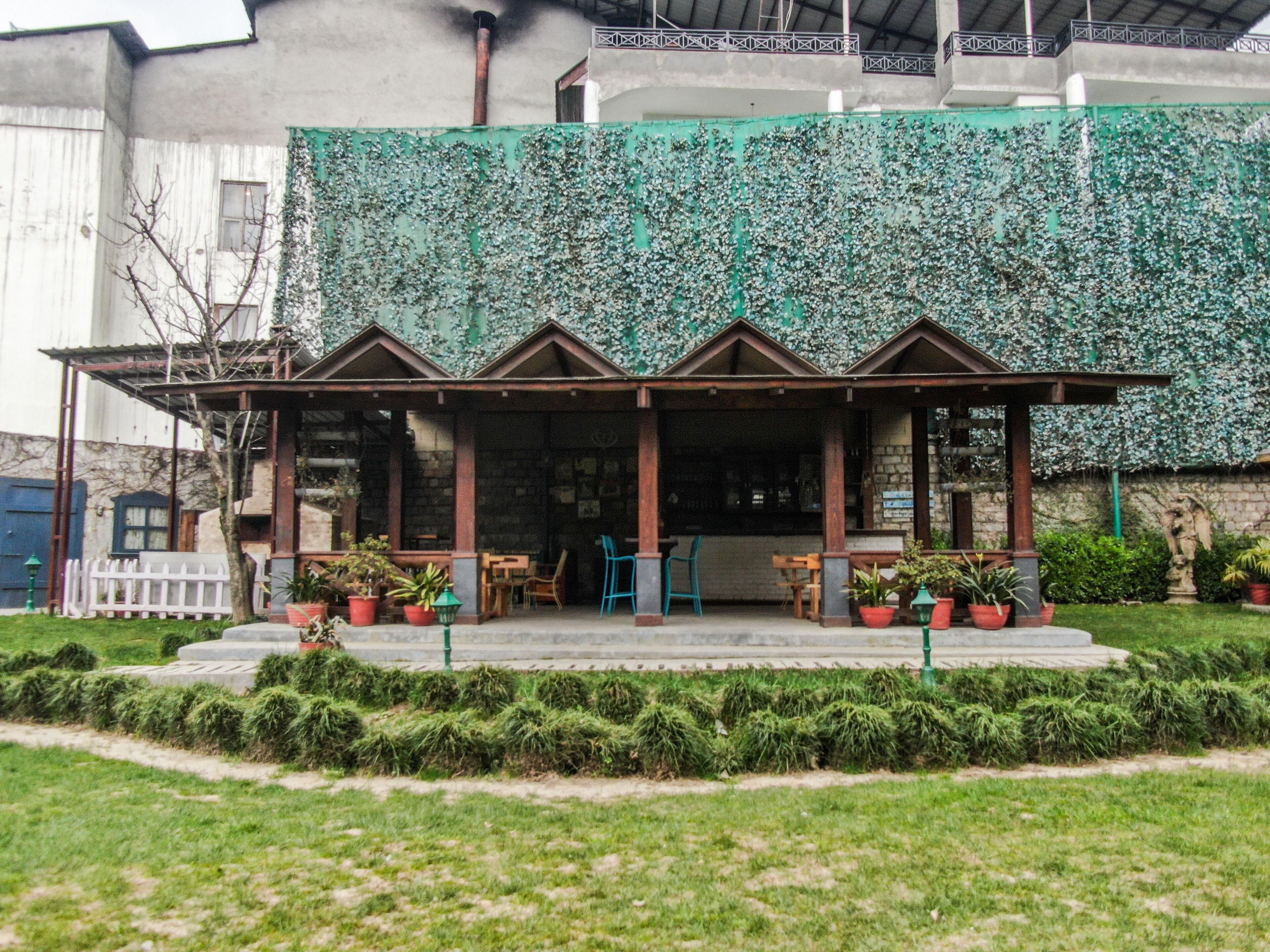 A building with a green ivy-covered wall, a covered outdoor seating area with tables and chairs, and a white fence in the foreground.
