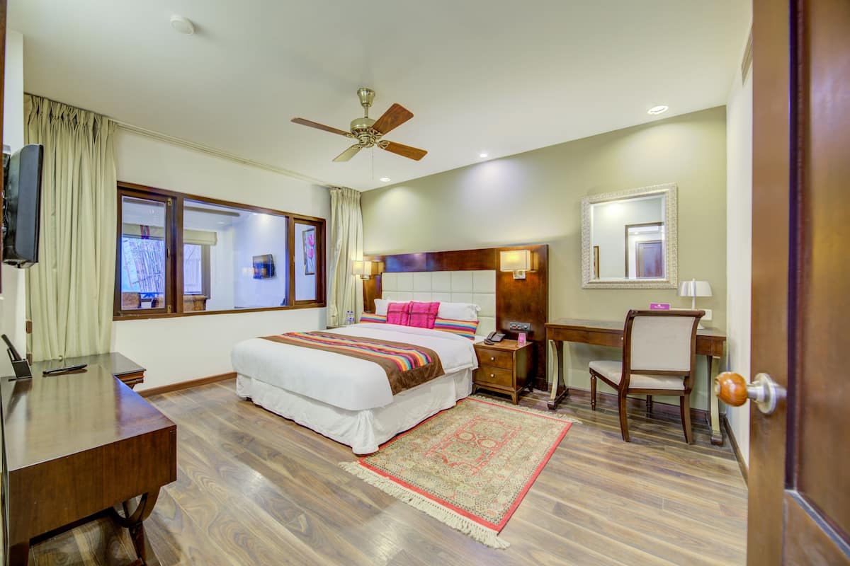 Une chambre d’hôtel avec un grand lit, un bureau, une chaise, un ventilateur de plafond et une fenêtre avec des rideaux.