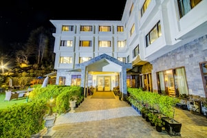 Un hotel de varios pisos con entrada central, sendero empedrado y plantas en macetas.