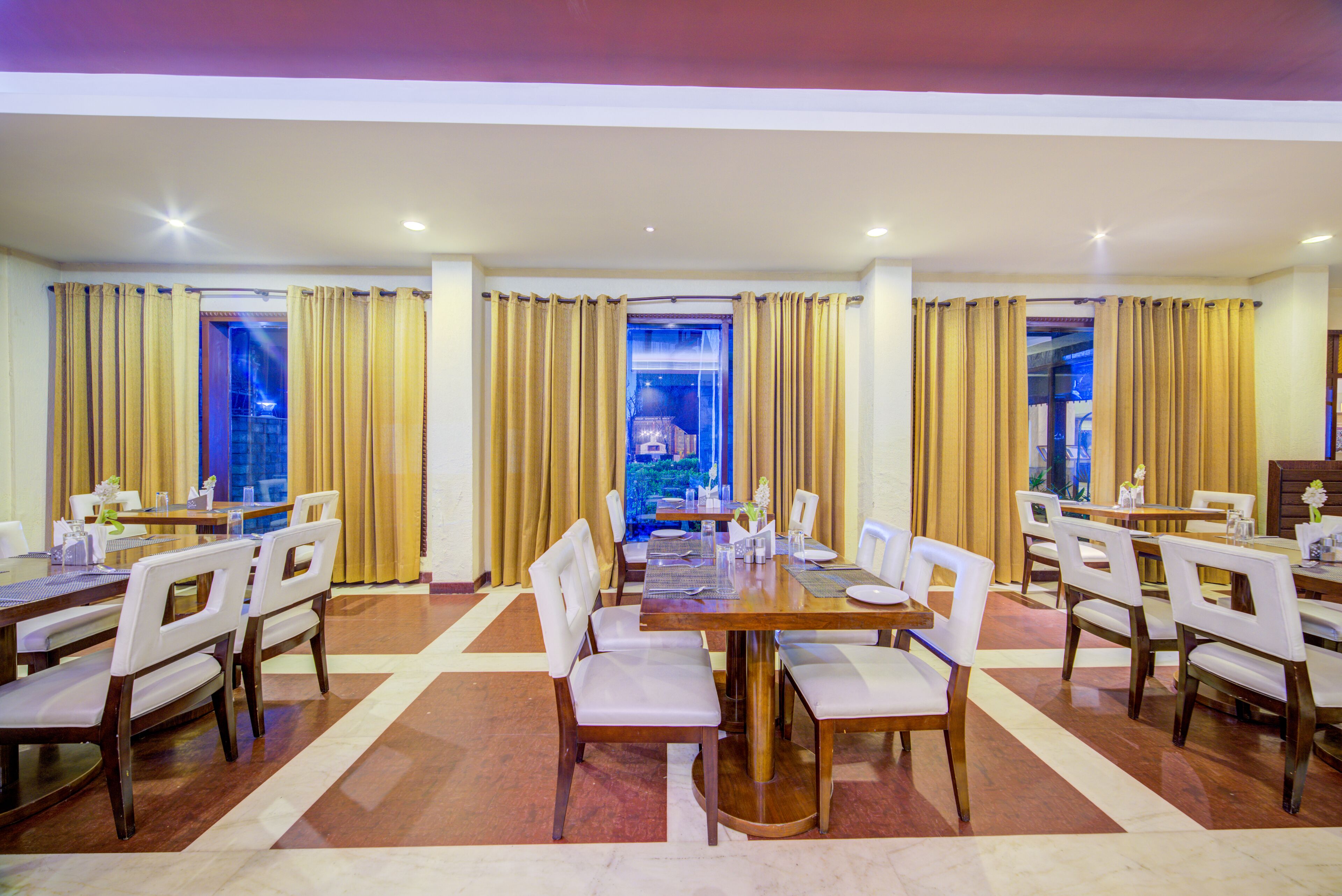 A spacious dining area with multiple tables and chairs, large windows with curtains, and a view of a garden.