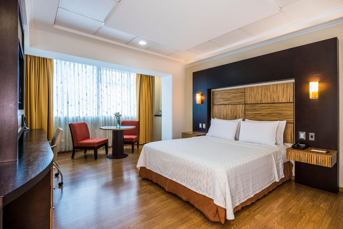 Hotelkamer met een groot bed, een bureau, twee stoelen, een klein tafeltje met een vaas en een raam met gordijnen.