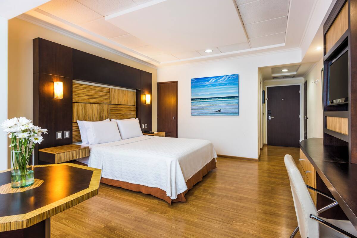Een hotelkamer met een bed, nachtkastje en een schilderij van een strandgezicht.