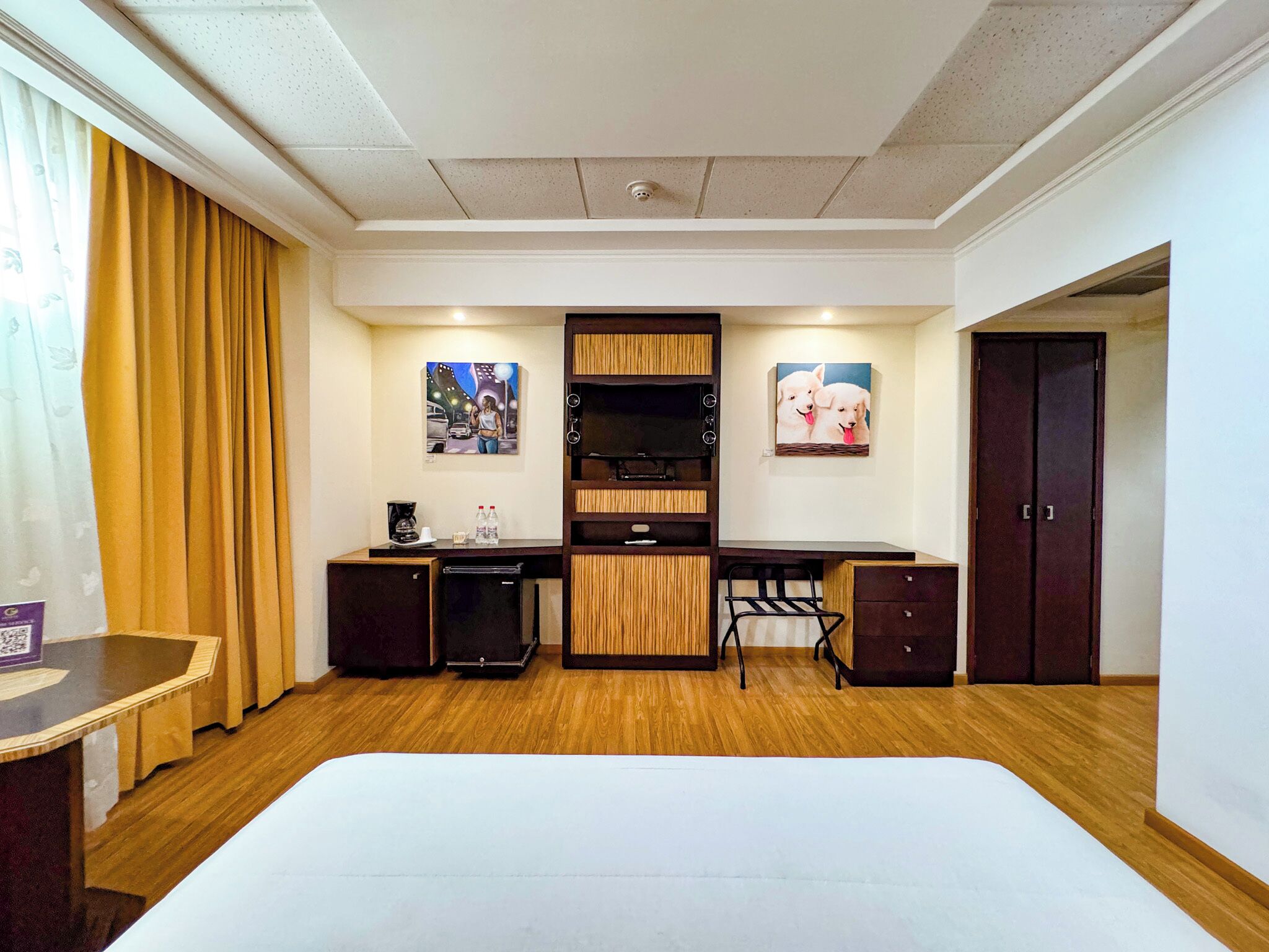 A hotel room with a bed, desk, and kitchenette. There are two paintings on the wall, a door, and a window with curtains.