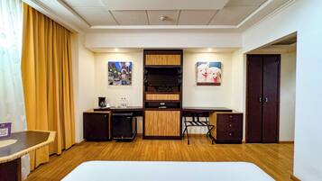 A hotel room with a bed, desk, and kitchenette. There are two paintings on the wall, a door, and a window with curtains.
