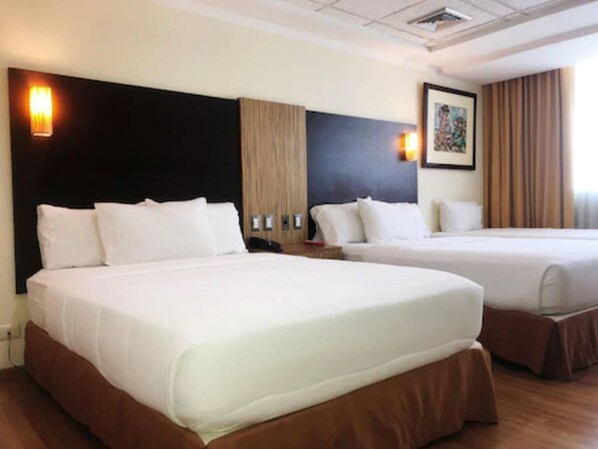 A hotel room with two beds, a dark headboard, a wooden accent wall, a framed picture, and a window with curtains.