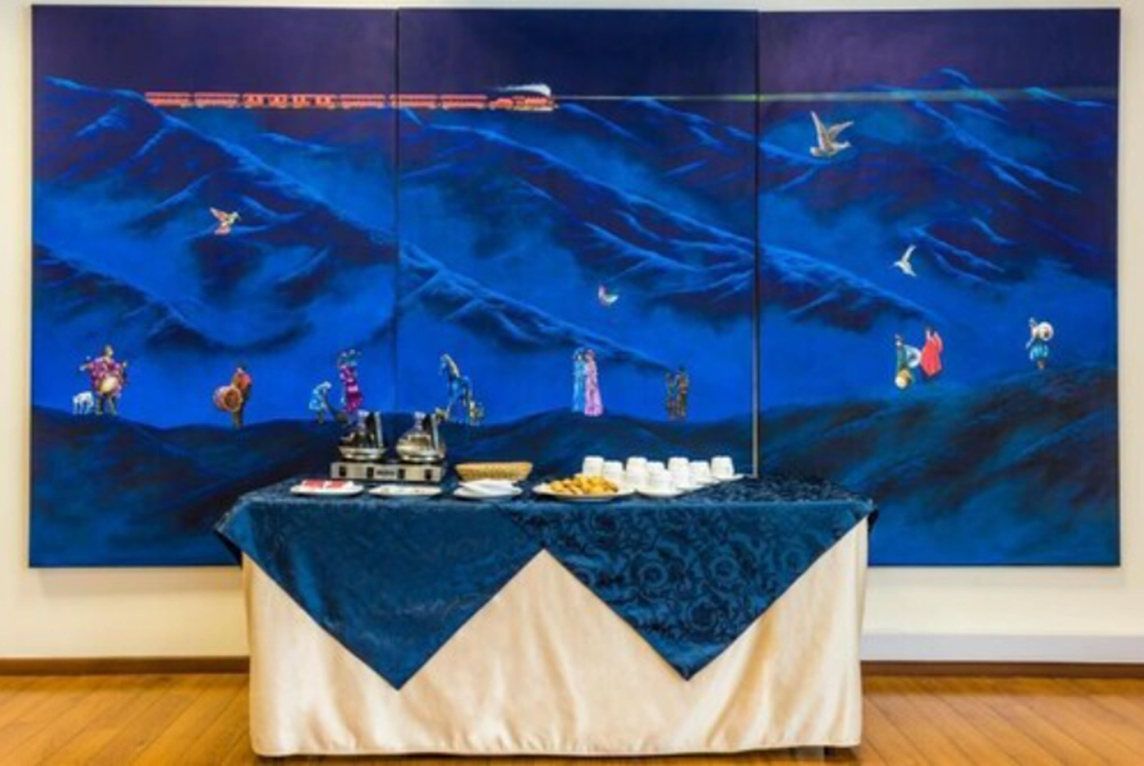 Una habitación con un cuadro grande que muestra un paisaje marino azul en la pared, una mesa con mantel y varios objetos sobre ella.