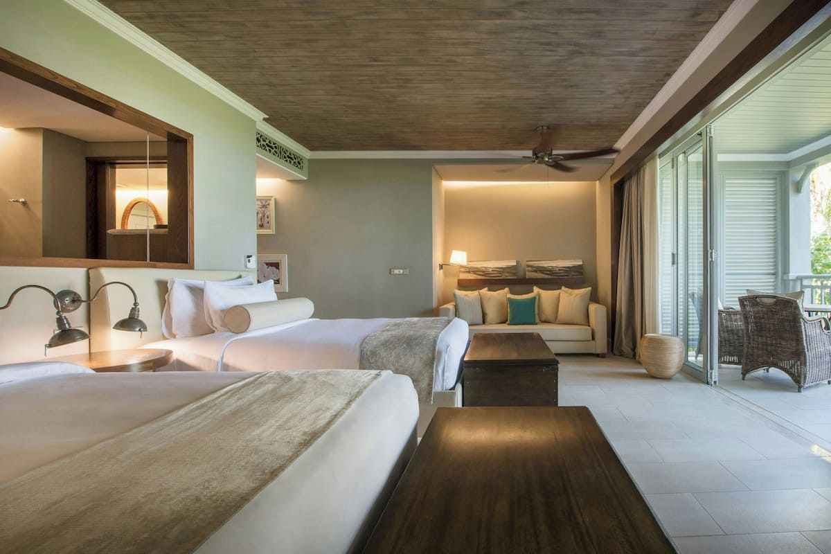 A hotel room with two beds, a sofa, a dining table, and a large window with curtains.