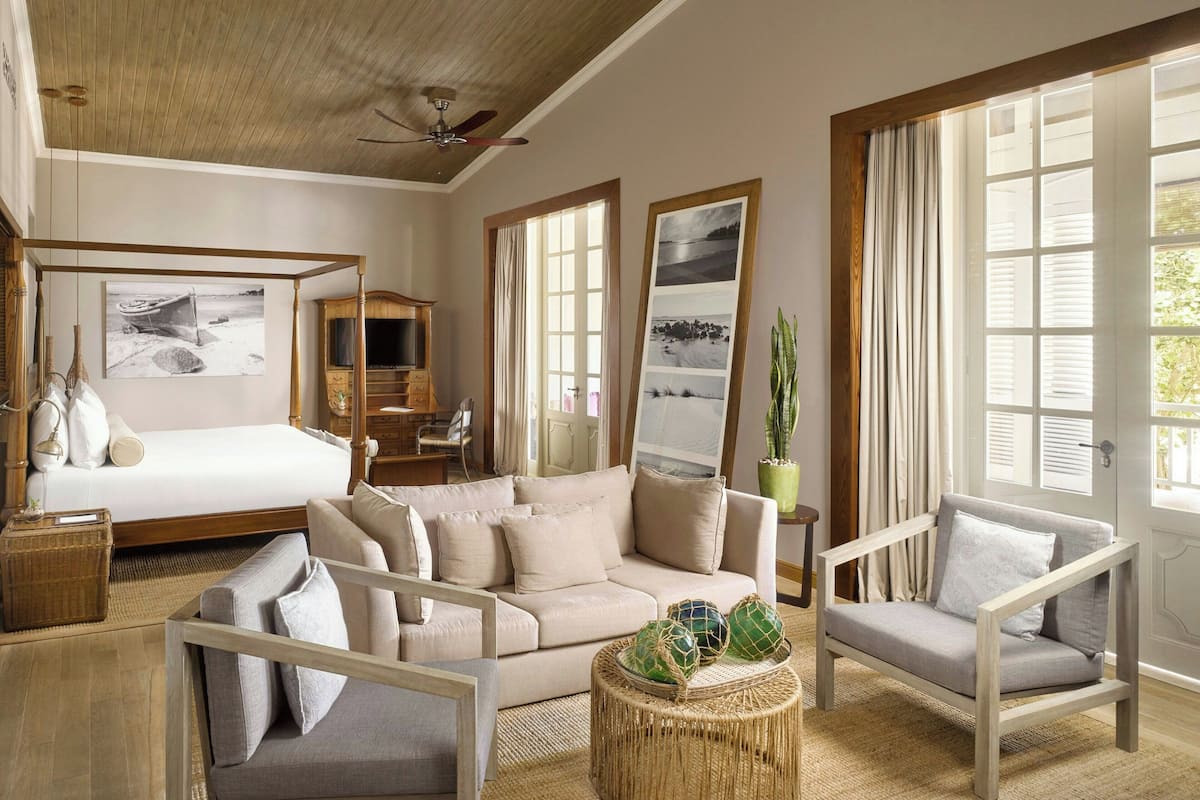 A spacious room with a bed, a sofa, a chair, a small table with decorative items, and large windows with curtains.