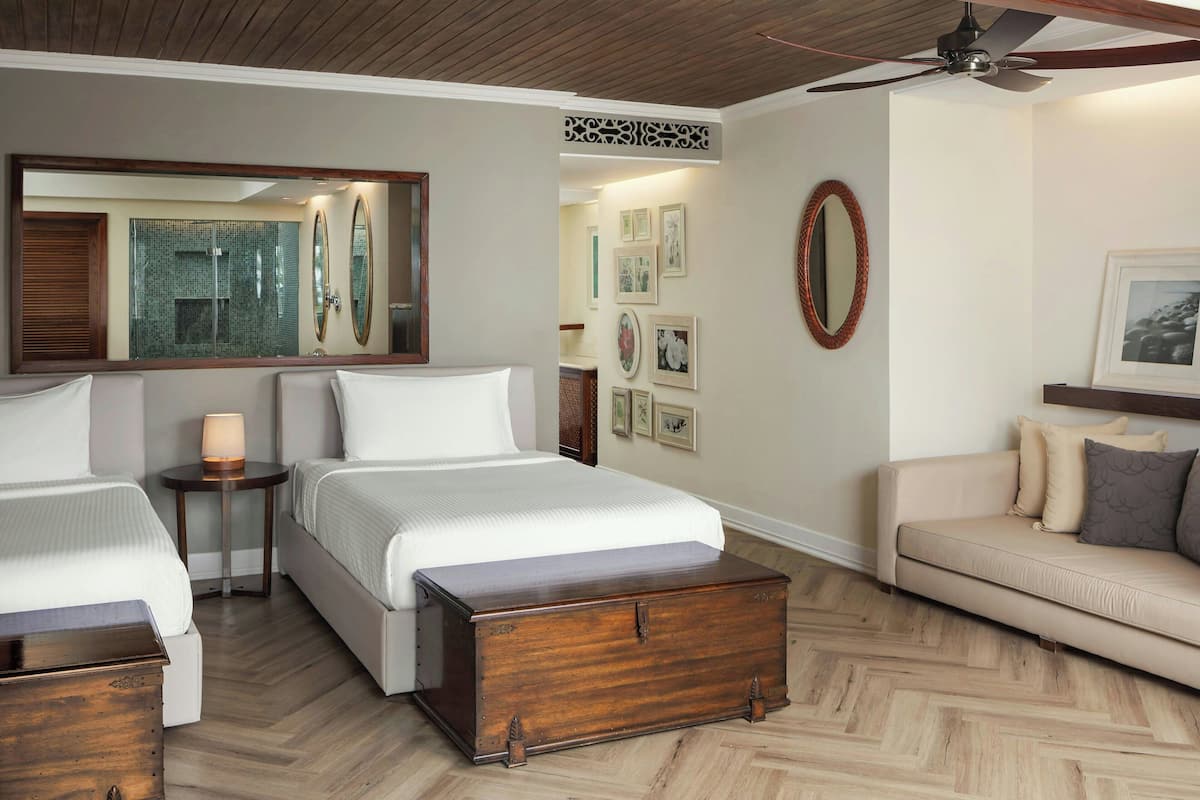 A hotel room with two beds, a wooden chest, a sofa, a ceiling fan, and framed pictures on the walls.