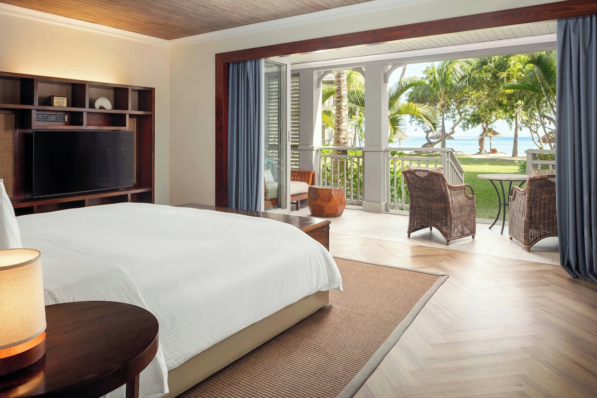 Habitación de hotel con una cama grande, televisión, balcón con mobiliario de mimbre y vista al océano.