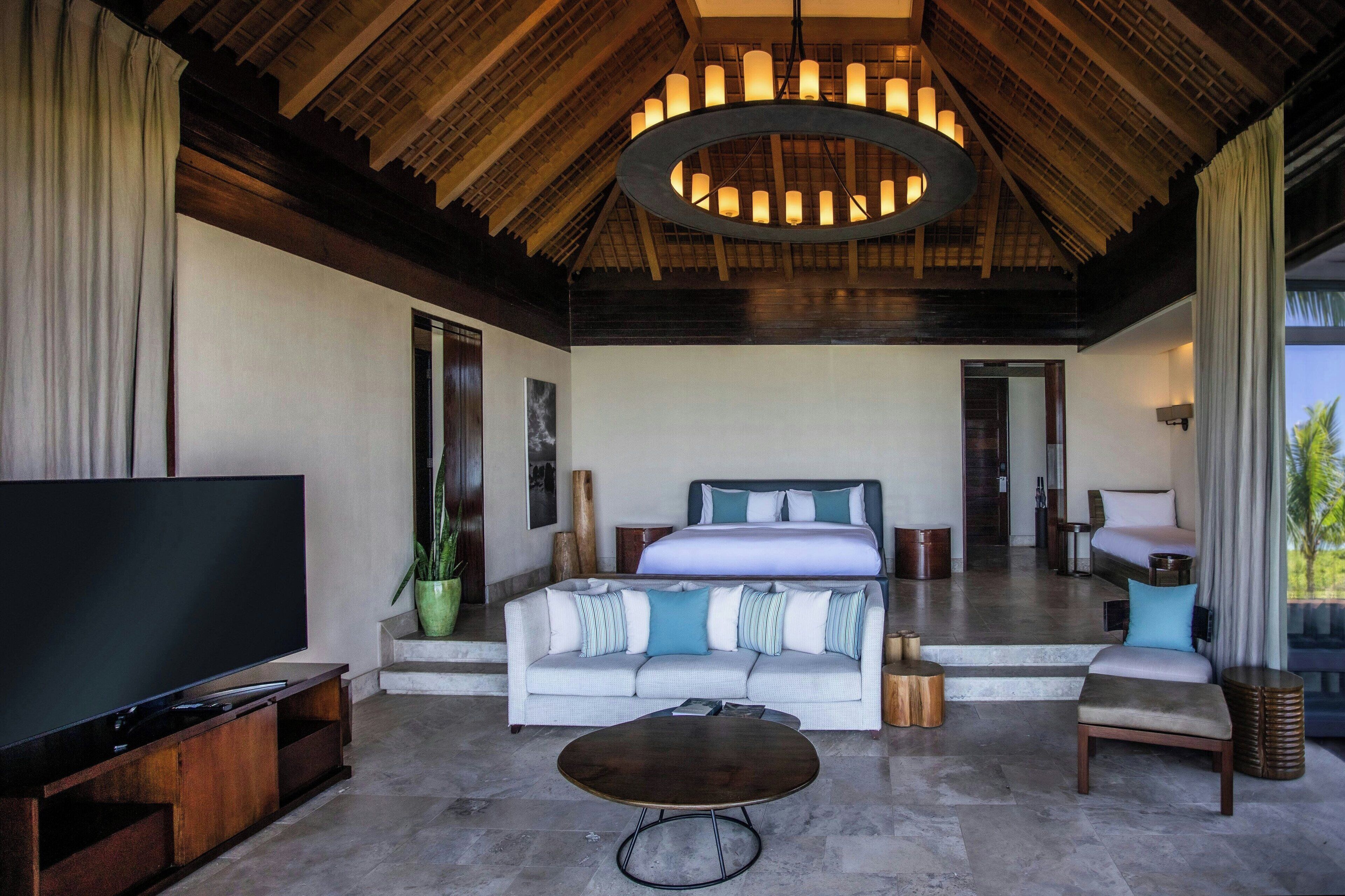 A spacious living room with a large flat-screen TV, a sofa with multiple cushions, a coffee table, and a ceiling chandelier.
