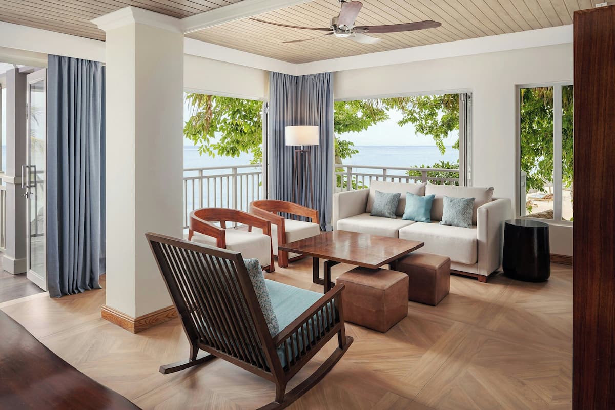 A spacious living area with a wooden floor, a sofa set, a rocking chair, and a table. Large windows offer a view of the ocean and greenery.