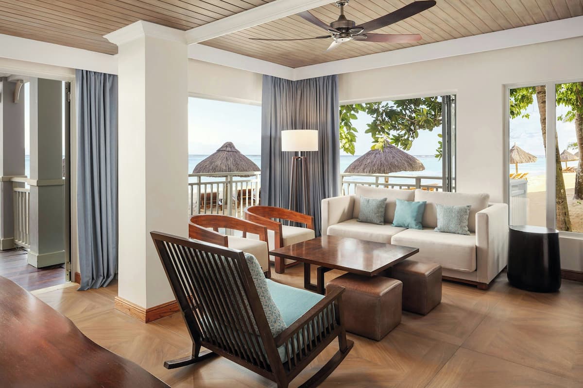 A spacious living area with a wooden ceiling, a ceiling fan, a sofa set, a coffee table, and a chair. There is a view of the beach through large windows.