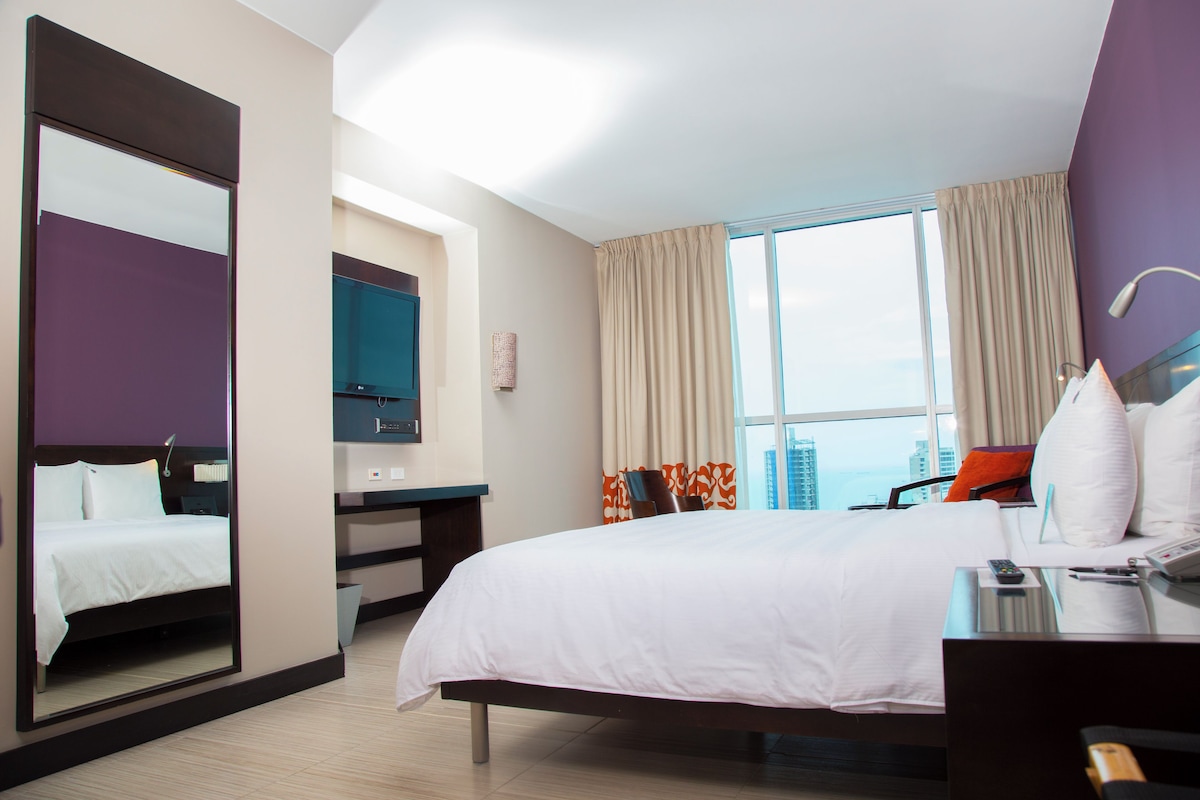 Una habitación de hotel con una cama grande, un escritorio, una silla, un espejo y una ventana con cortinas.