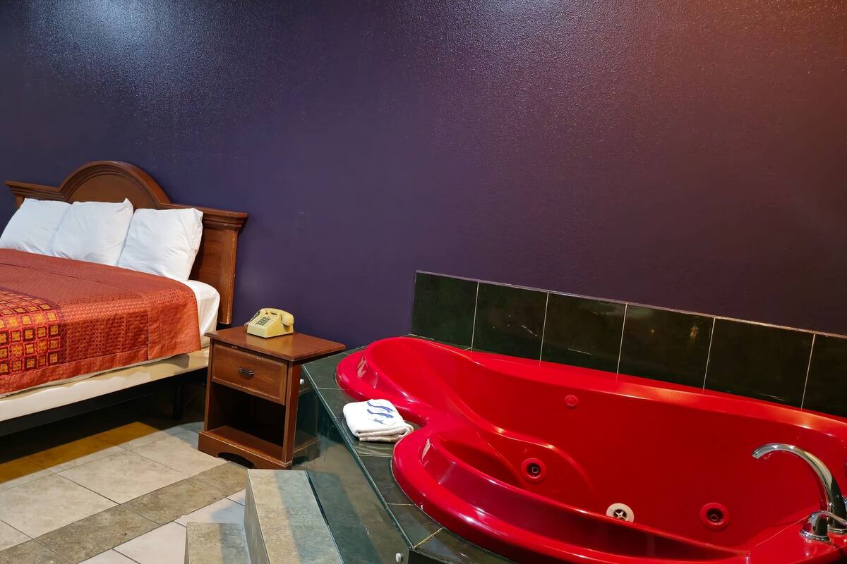 A hotel room with a bed, a bedside table with a telephone, a red bathtub, and a window with yellow curtains.
