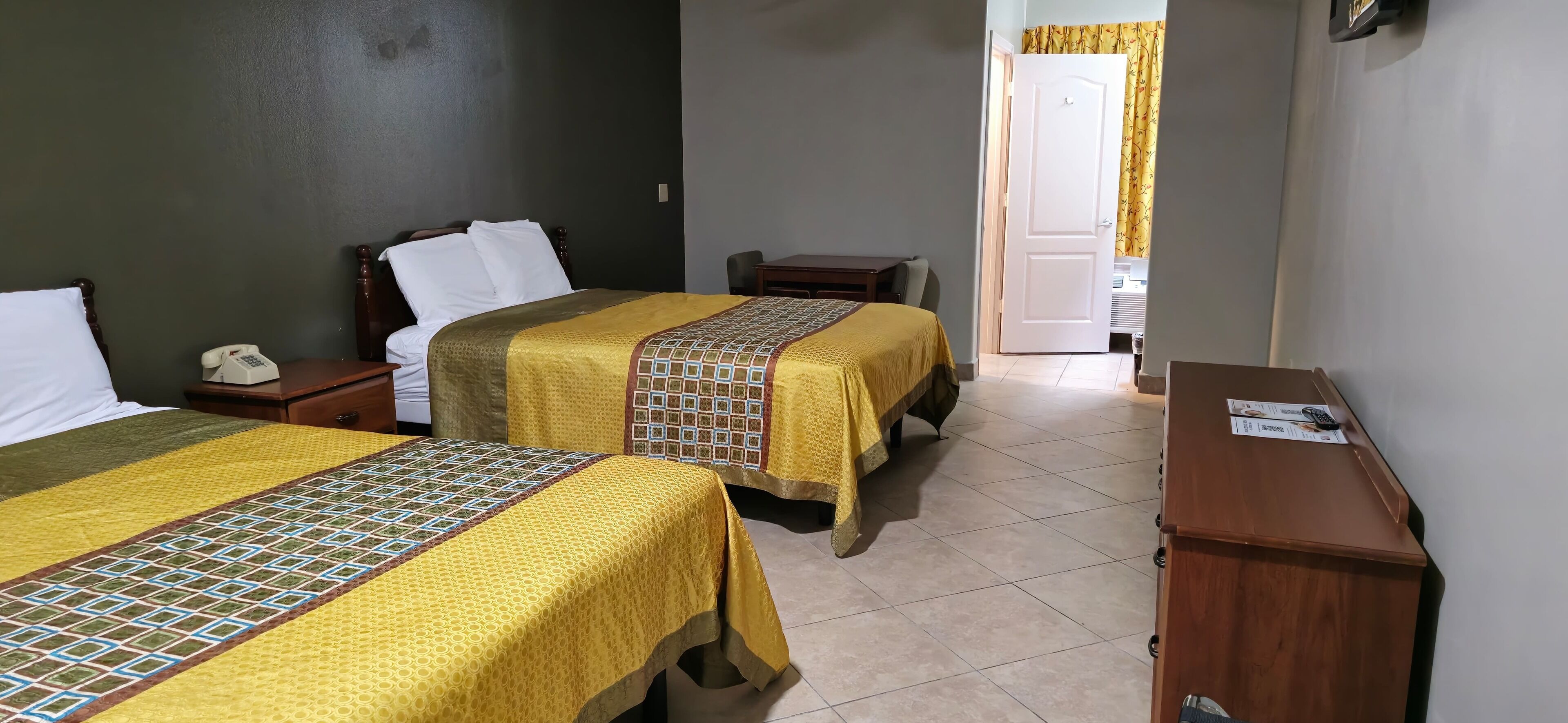 A hotel room with two beds, a wooden nightstand with a telephone, a desk with a computer and papers, and a door leading to a bathroom.