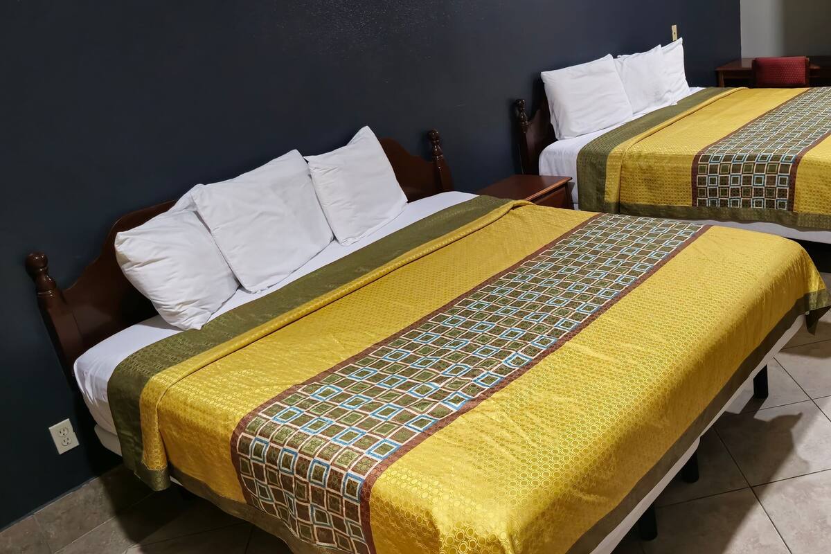 Two single beds with wooden frames, white pillows, and yellow and green patterned bedspreads. There is a small wooden table and a red chair in the room.