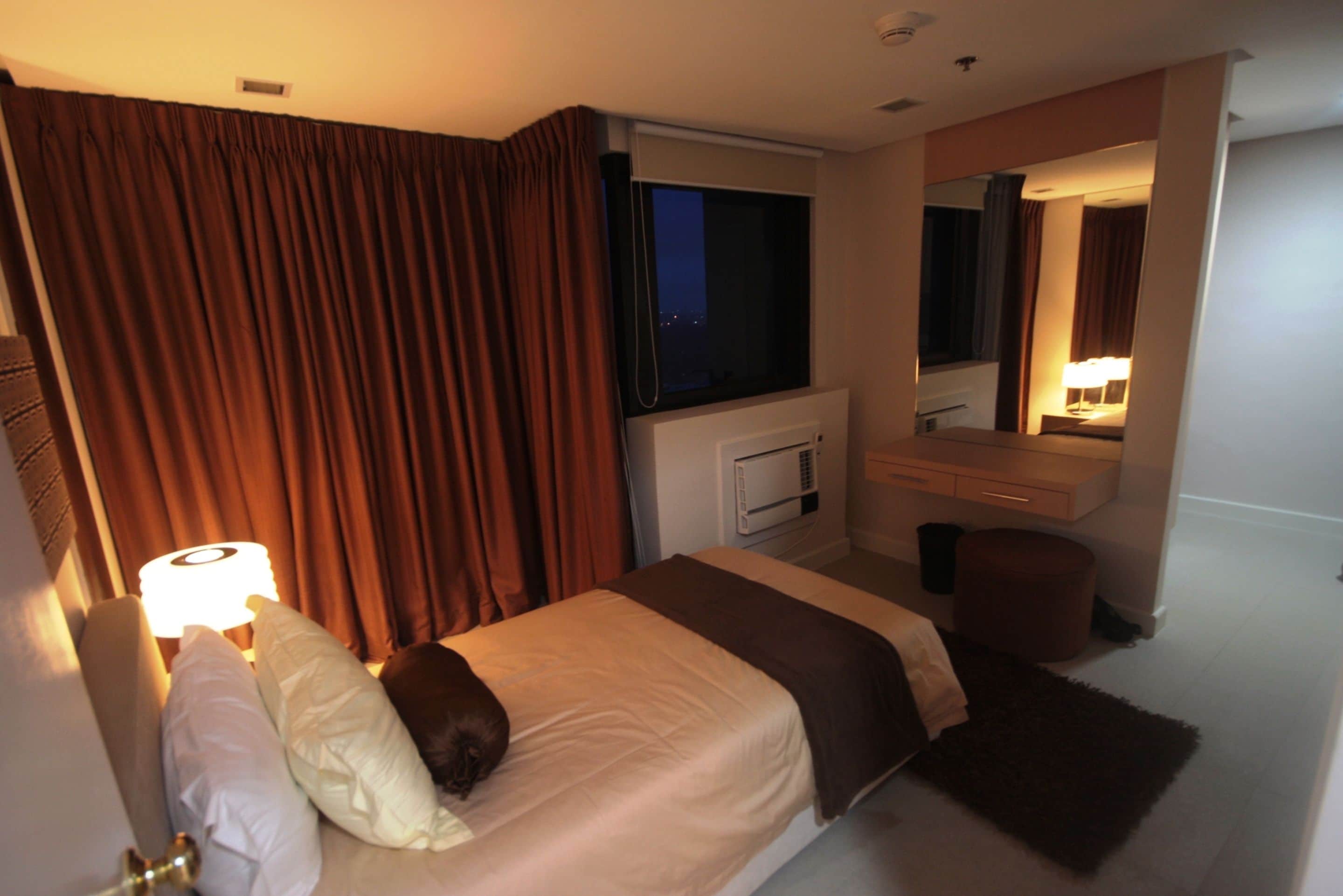 Een hotelkamer met een bed, een raam, een spiegel, een nachtkastje en een lamp.