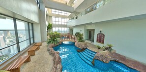Una piscina interior con vistas a la ciudad, bancos de madera y plantas en macetas.