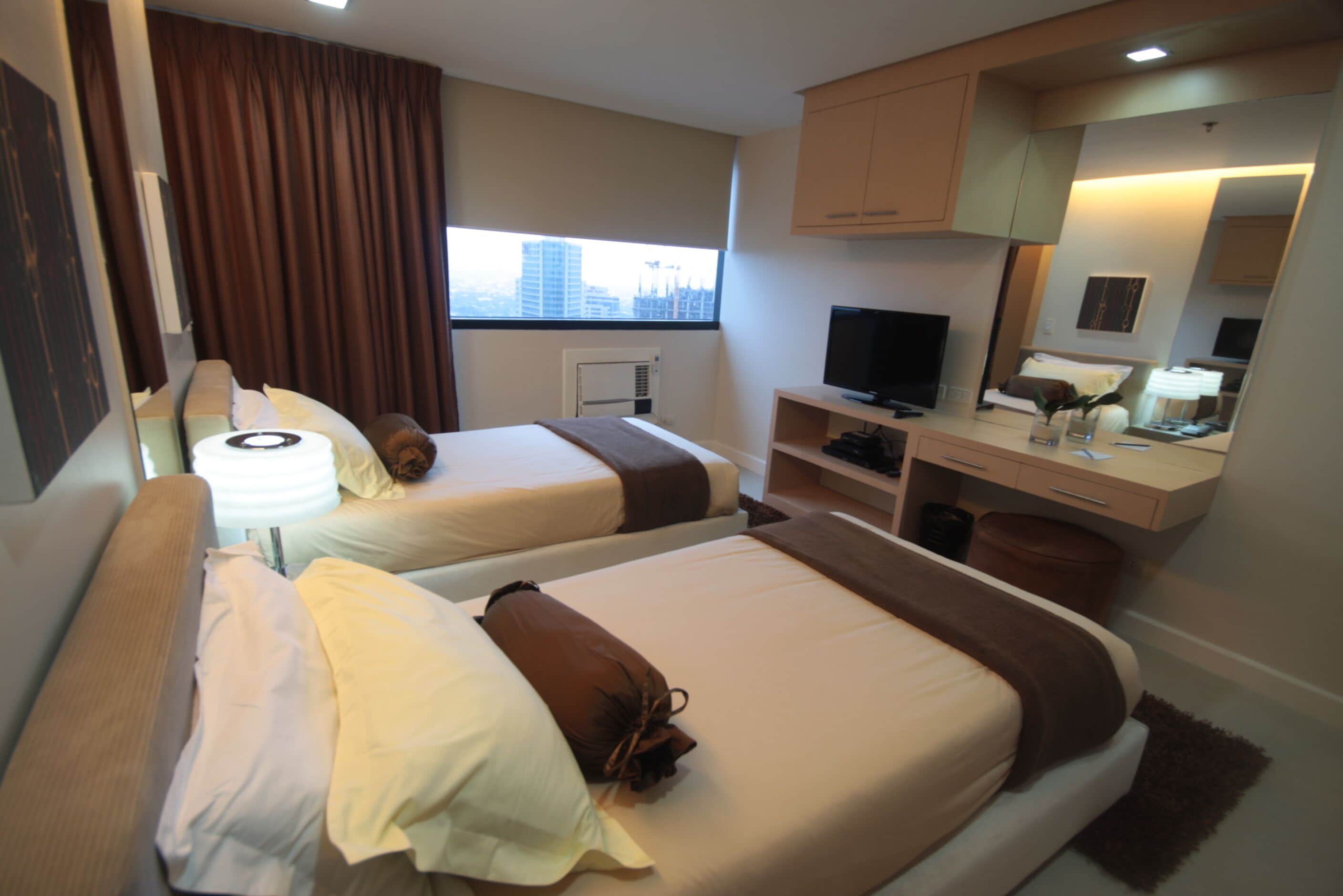 A hotel room with two beds, a TV, a desk, and a window with a city view.