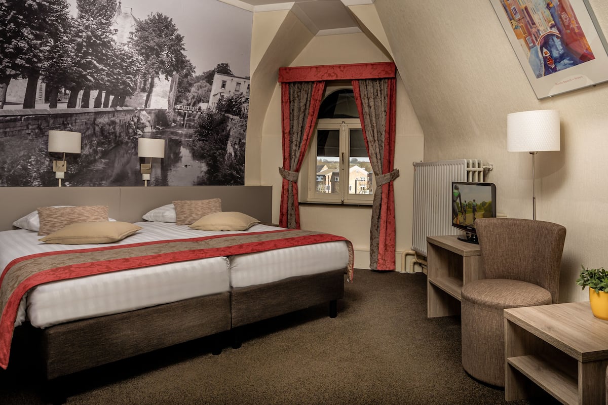 A bedroom with a large bed, a desk, a chair, a radiator, and a wall with a cityscape mural.