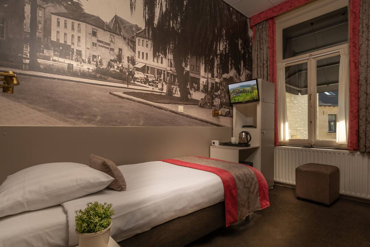 A hotel room with a bed, a small table with a kettle, a television, and a window with curtains.