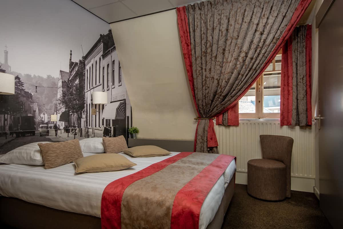 A hotel room with a large bed, a chair, a window with red curtains, and a mural of a street scene on the wall.