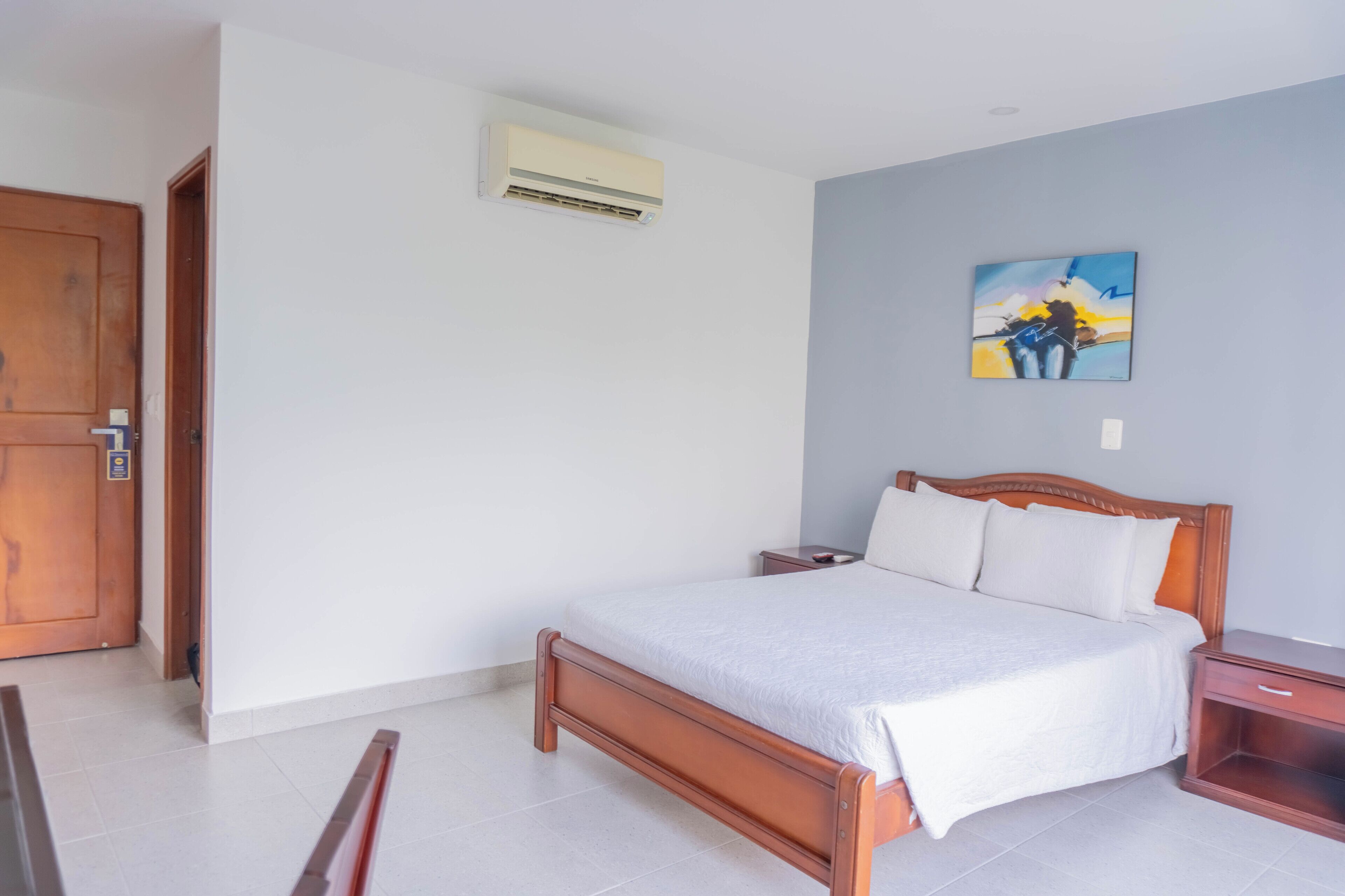 A bedroom with a wooden bed, a nightstand, a wall-mounted air conditioner, and a painting on the wall.