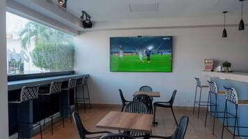 A modern bar area with a large TV screen displaying a soccer game, black bar stools, and a wooden table.