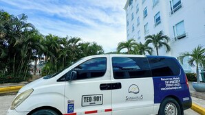 Uma carrinha branca e azul estacionada em frente a um hotel com palmeiras e um céu limpo ao fundo.