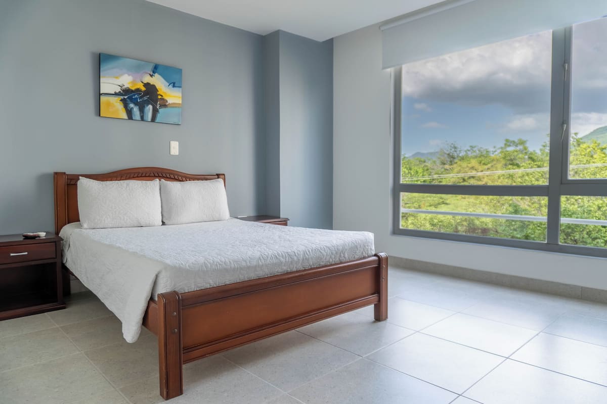 Un dormitorio con una cama de madera, una mesita de noche, un ventanal con vistas a la vegetación y un cuadro en la pared.