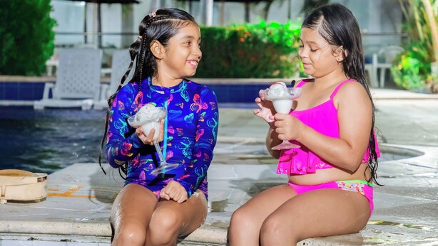 Duas pessoas sentadas à beira de uma piscina em cenário tropical, com palmeiras e um edifício ao fundo.