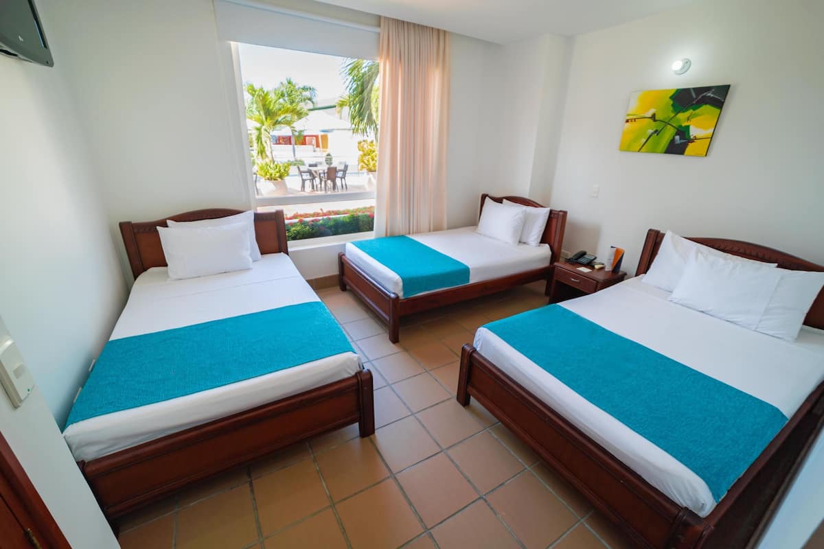 Habitación de hotel con tres camas de madera, cada una con ropa de cama blanca y detalles azules. Se ve una ventana con vistas al exterior, incluyendo palmeras y un edificio.
