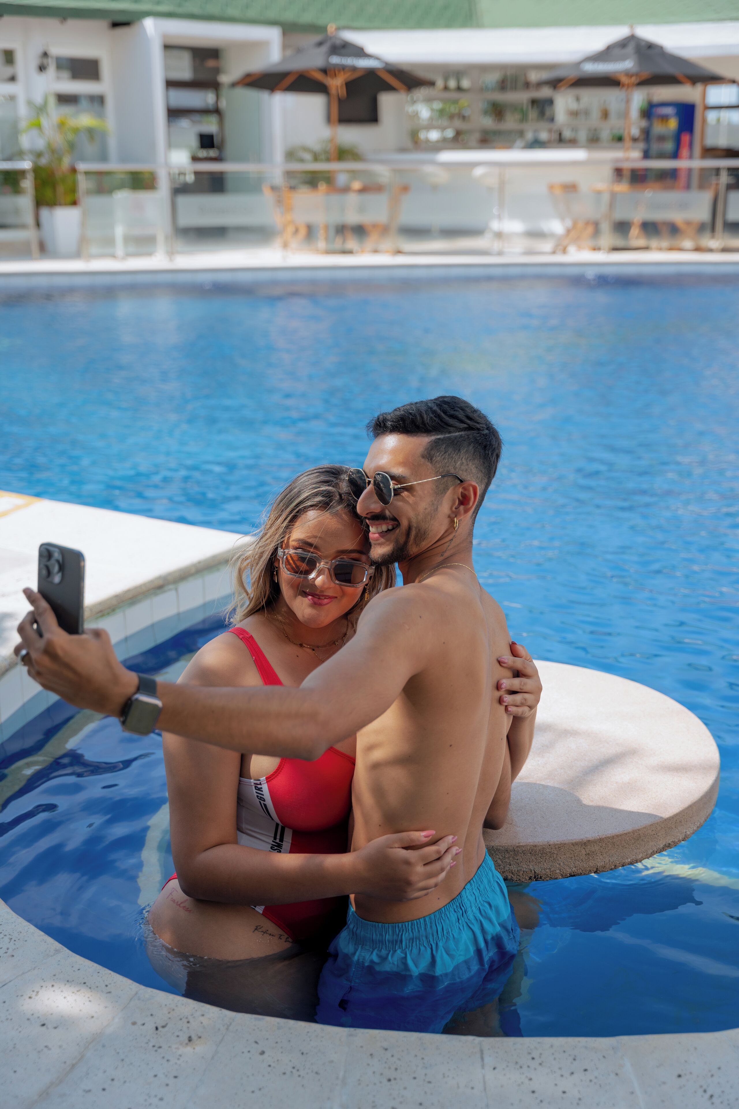 A couple taking a selfie by a pool with a bar and lounge area in the background.