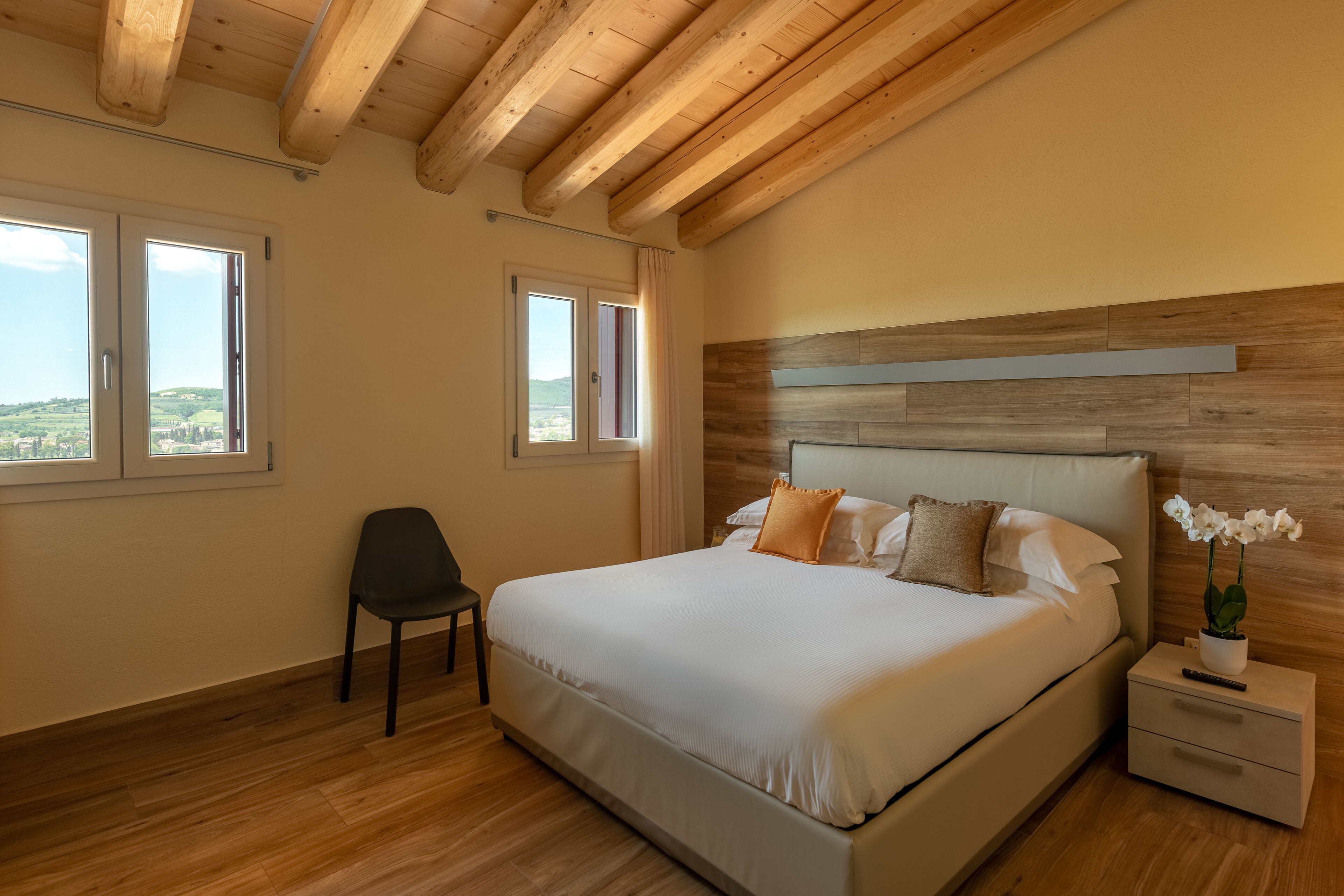 A bedroom with a large bed, wooden ceiling beams, a nightstand with a remote, and a chair.