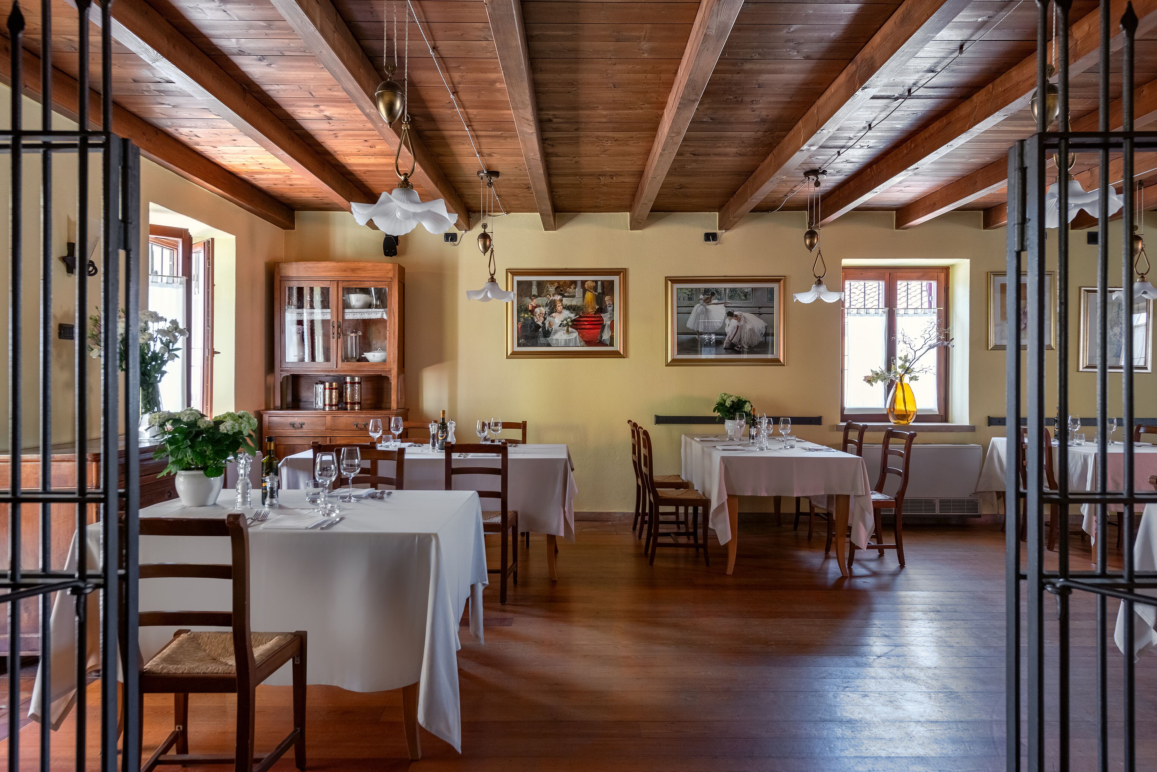 Un espace repas agrémenté de poutres en bois, de tables et de chaises blanches. Les murs sont ornés de tableaux et on y trouve une vitrine aux portes en verre.