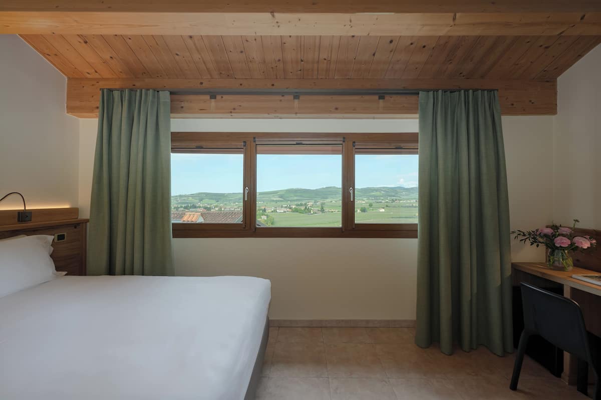 A bedroom with a bed, a desk, a chair, a window with curtains, and a view of the countryside.