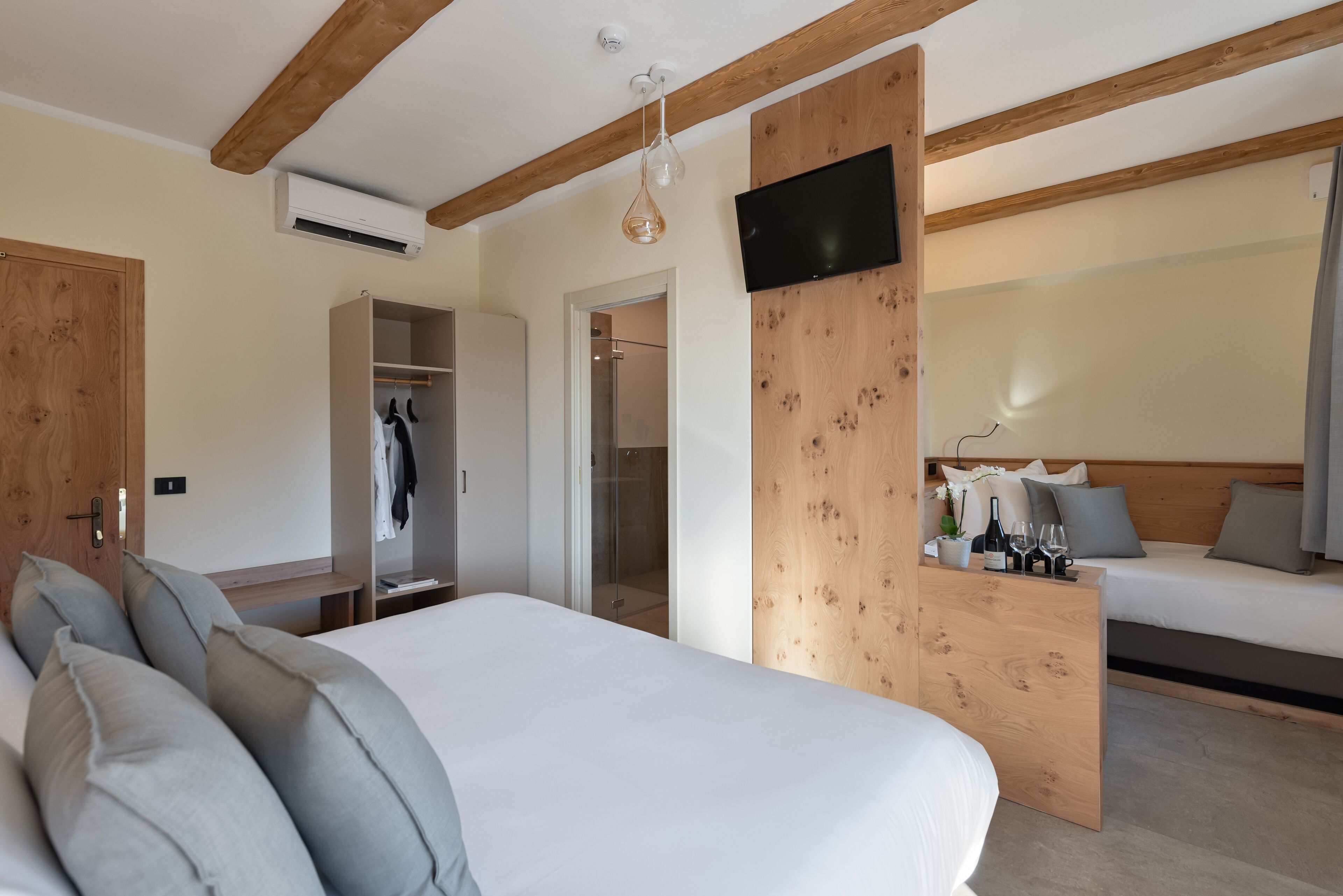 Une chambre avec un plafond en bois, un lit avec des oreillers gris, une table de chevet avec une bouteille et des verres, et un téléviseur fixé au mur.