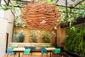 Una cesta colgante de mimbre en una cafetería con sillas verdes y una pared de ladrillo.