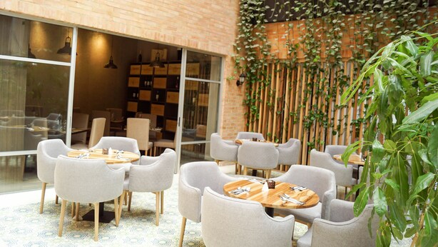 Un comedor moderno con mesas redondas y sillas grises, una pared de ladrillo y un muro cubierto de plantas de bambú.