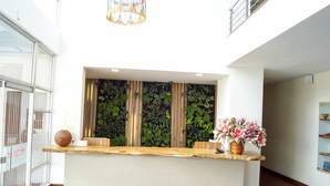 Un mostrador de recepción con encimera de madera, un ventanal con persianas verticales y una pared verde con plantas.