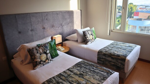 A hotel room with two beds, each with a grey headboard and white bedding. There is a small bedside table with a lamp and a phone. The room has a large window with a view of buildings and trees.